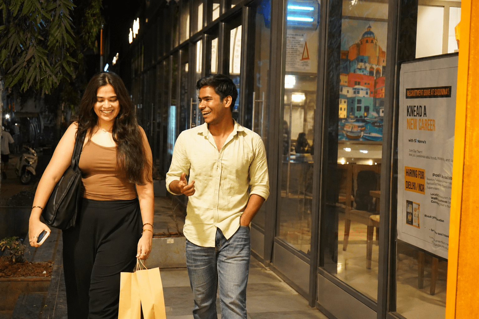 Two friends laughing while walking together past a cafe on a city street at night