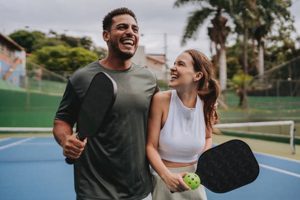 Two people laughing on an outdoor court after a game, each holding a paddle